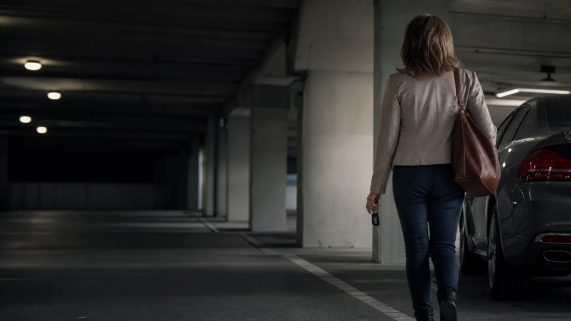 A woman in a light jacket and jeans, carrying a brown shoulder bag and holding keys, walks away from the camera toward a parked car in a dim concrete parking garage.