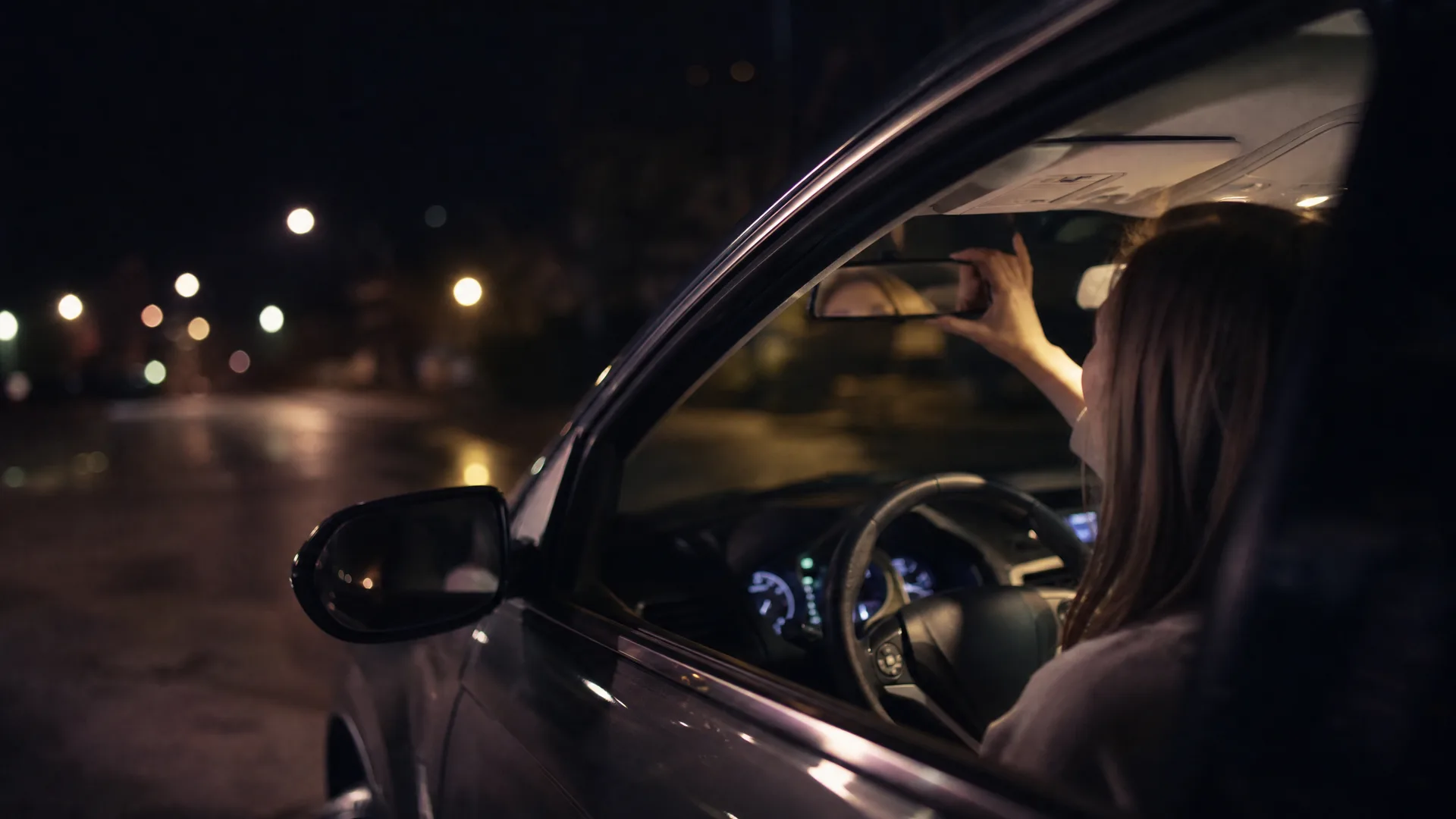A woman sitting in the driver's seat of a car at night reaches up to adjust the rearview mirror, seen through the open passenger-side window with blurred street lights behind.